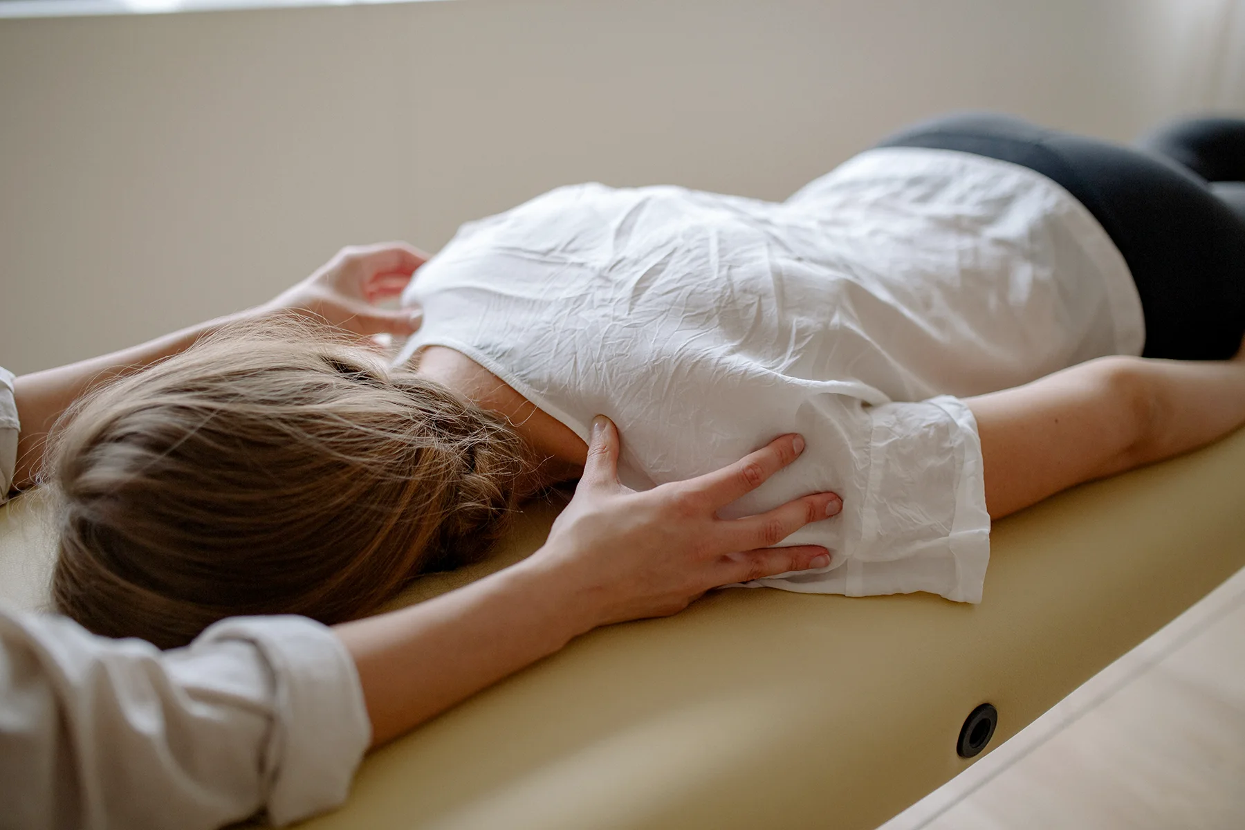 Die Physiotherapeutin übt sanften Druck auf die Schultern einer auf der Therapieliege liegende Patientin aus.